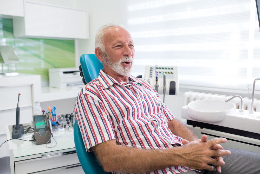 An older man with a white beard sits in a dentist’s chair, wearing a striped shirt. He smiles slightly with hands folded. Dental tools and equipment are visible in the bright, modern clinic background.