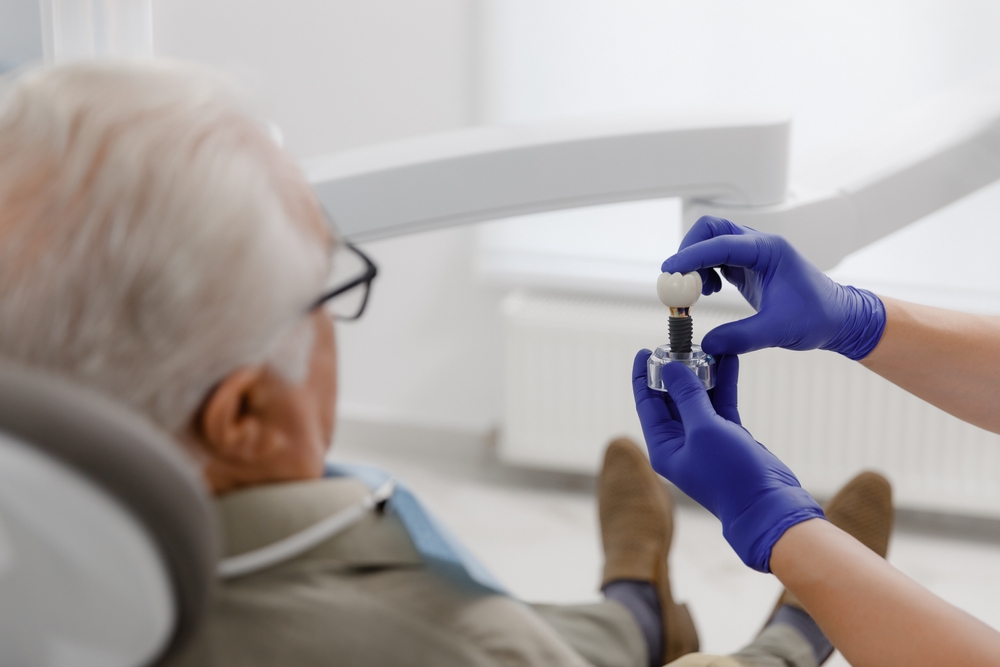 A dentist in blue gloves shows a dental implant model to an older man with gray hair, seated in a dental chair in a clinical setting.